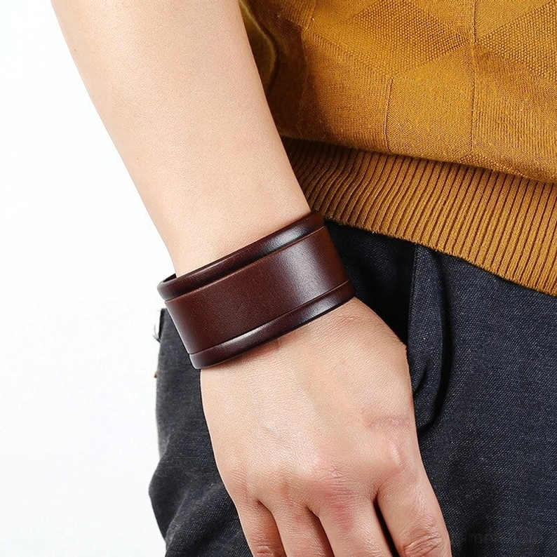 Breites braunes Leder-Gothic-Armband auf Handgelenk getragen, Lifestyle-Ansicht mit gelbem Strickpullover und dunkler Hose, zeigt natürliche Trageposition und Styling-Effekt