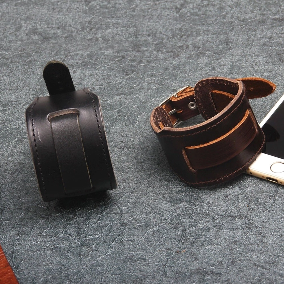 Zwei breite Leder Gothic Armbänder in Schwarz und Braun auf dunkler Steinoberfläche präsentiert mit Smartphone als Größenreferenz, verstellbare Schnallenverschlüsse deutlich sichtbar