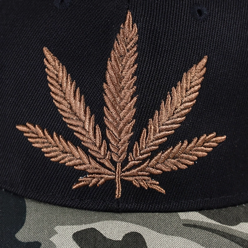 Detailaufnahme der braunen Hanfblatt-Stickerei auf Gothic Kappe mit dreidimensionaler Textur, Camouflage-Schirm-Kantenstruktur, hochwertige Näh-Details und botanische Motiv-Verarbeitung