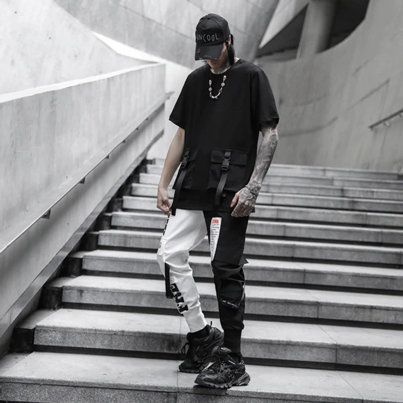 Ganzansicht eines männlichen Modells mit tätowierten Armen, Baseballkappe und schwarzem T-Shirt, das die schwarze und weiße Gothic-Patchwork-Cargo-Hose mit klobigen Sneakern auf Betontreppe trägt