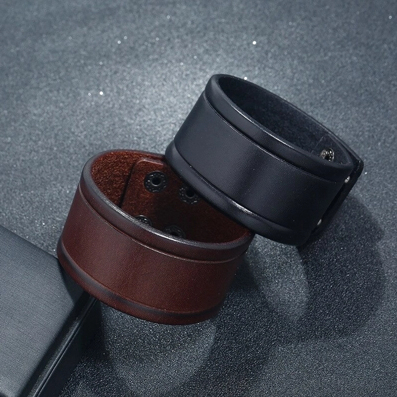Schwarzes und braunes breites Leder-Gothic-Armband im Vergleich, Flat-Lay-Komposition auf dunklem Hintergrund, zeigt beide Farbvarianten und deren unterschiedliche Charakteristiken
